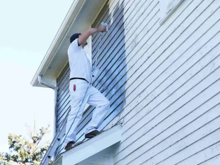 Painter working on ladder to paint white exterior siding and window trim