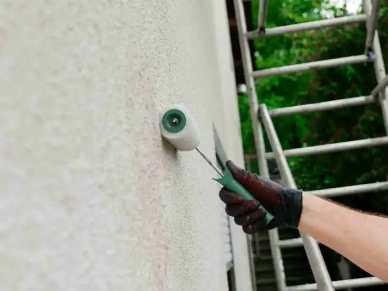 Gloved hand using a small roller to apply light paint on an exterior wall